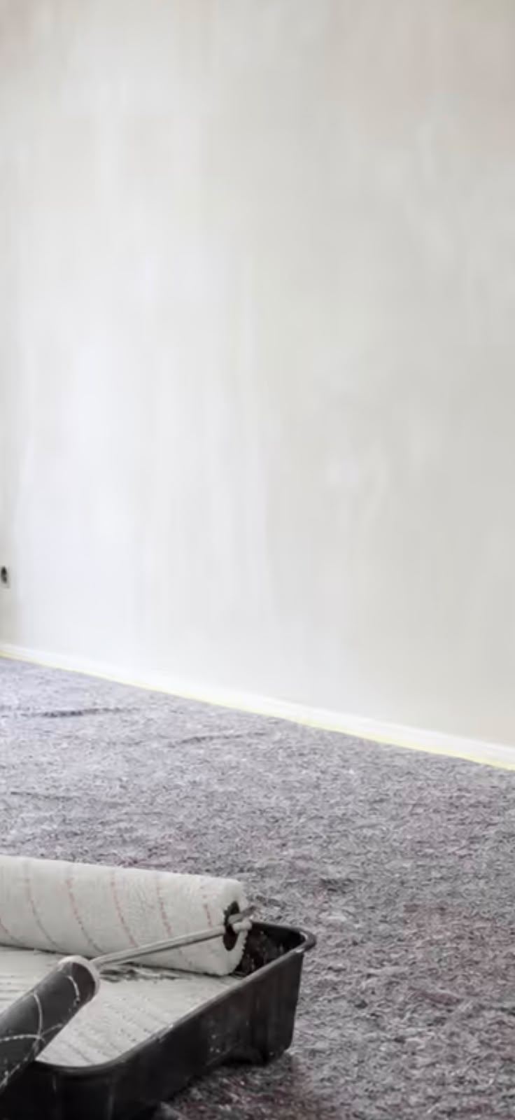Hochformatiges Detail einer frisch gestrichenen hellen Wand mit abgeklebter Fußleiste und Farbrolle in der Farbwanne auf dem Abdeckvlies.