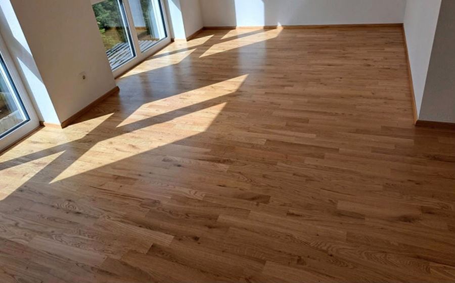Fertig verlegter Eichenparkettboden in einem hellen leeren Raum, von der Sonne durch die bodentiefen Fenster beschienen.