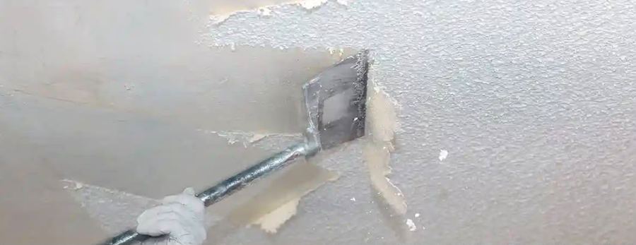 Behandschuhte Hand löst mit einem Spachtel alte Beschichtung von einer Decke ab, sehr breites Banner-Format.