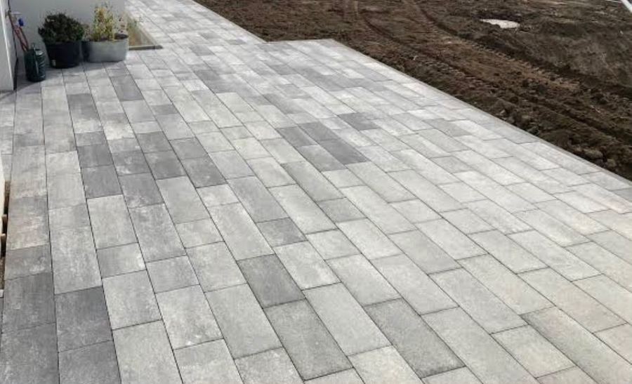 Fertig verlegte Fläche aus grau melierten Betonplatten entlang einer Hauswand, angrenzend noch offenes Erdreich.