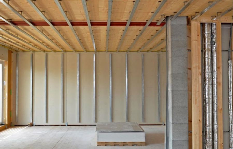 Beplankte Trockenbauwand mit sichtbarem Metallständerwerk unter einer Holzbalkendecke im Rohbau, davor ein Stapel Gipskartonplatten auf Paletten.