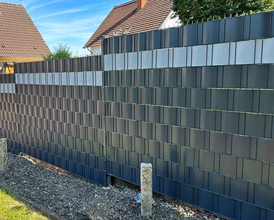 Anthrazitfarbener Doppelstabmattenzaun mit eingezogenen Sichtschutzstreifen auf Kiesstreifen bei Sonnenschein.