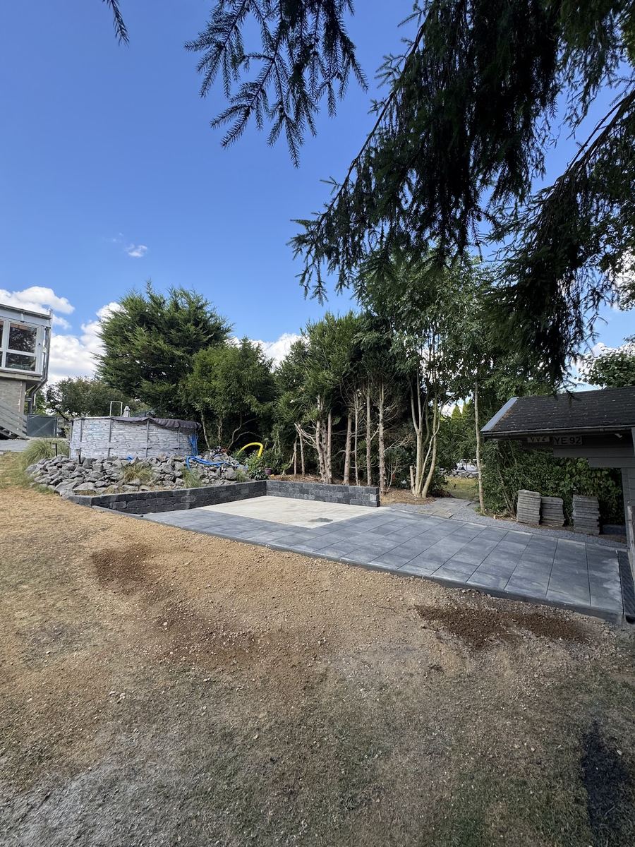 Fertig angelegte Pflasterfläche mit Betonfundament, niedriger Blocksteinmauer und Natursteinböschung im Garten.
