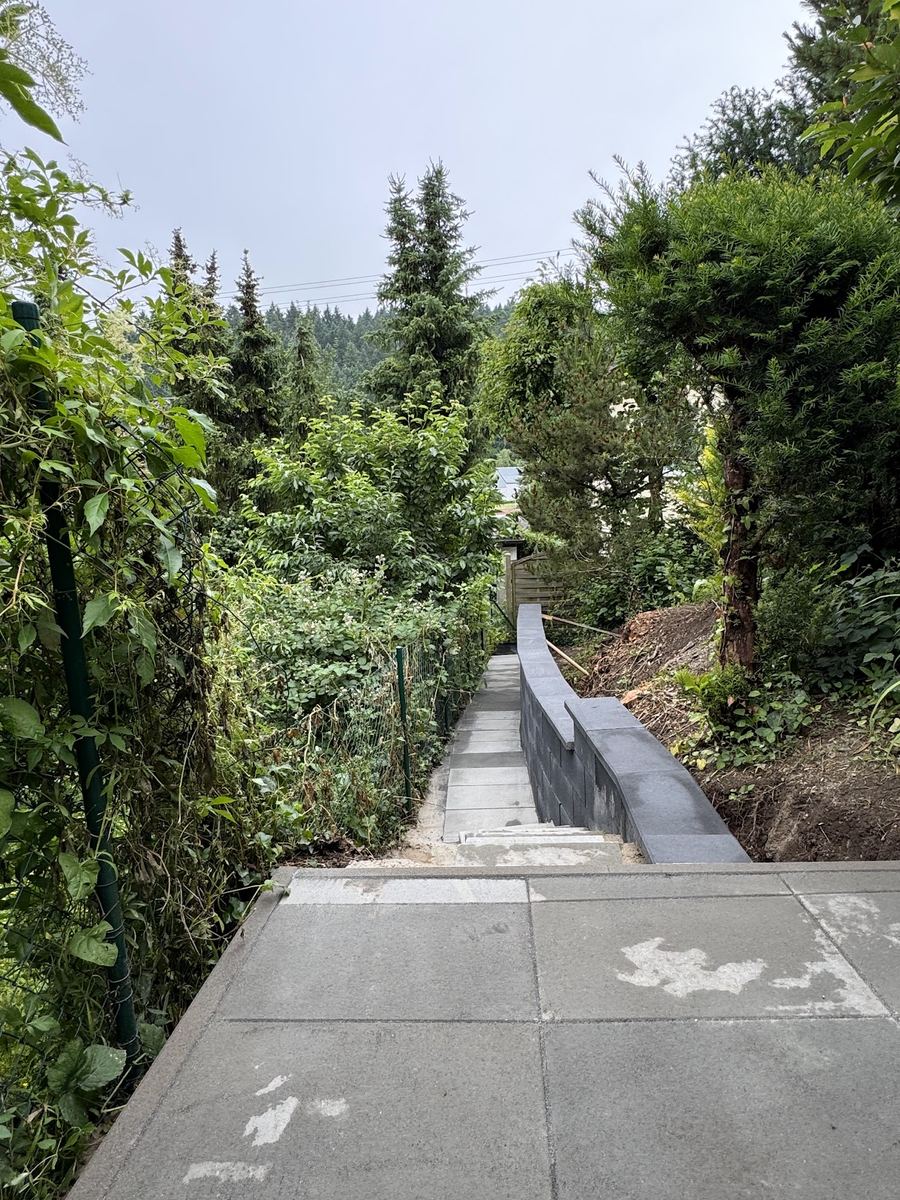 Neu gebauter Gartenweg mit Plattenbelag und anthrazitfarbener Stützmauer am Hang, Erdarbeiten noch sichtbar.