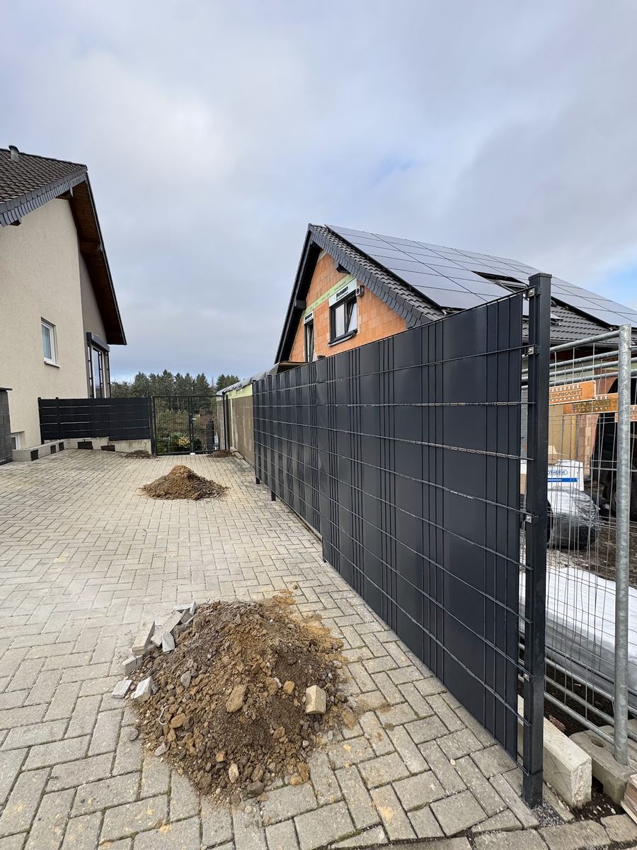 Neu montierter anthrazitfarbener Doppelstabmattenzaun mit Sichtschutzstreifen an einer gepflasterten Einfahrt, Baustelle im Hintergrund.
